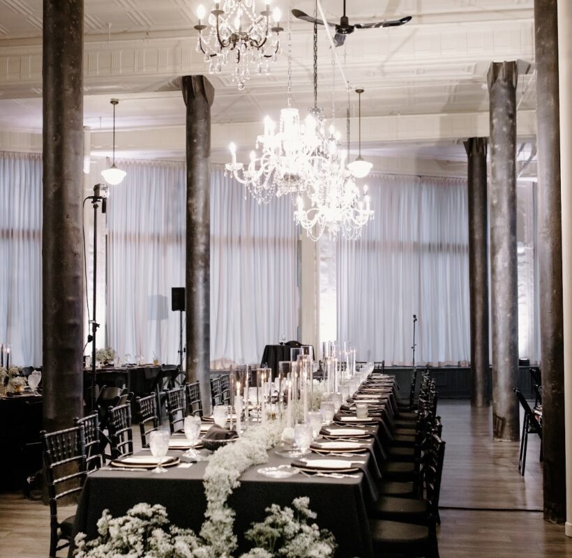 Long rectangle table decorated with black table cloth, glasses, and white flowers, in a room with chandeliers and black concrete pillars