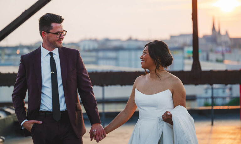 bride holding her wedding dress train and groom in a maroon suit holding hands on a rooftop