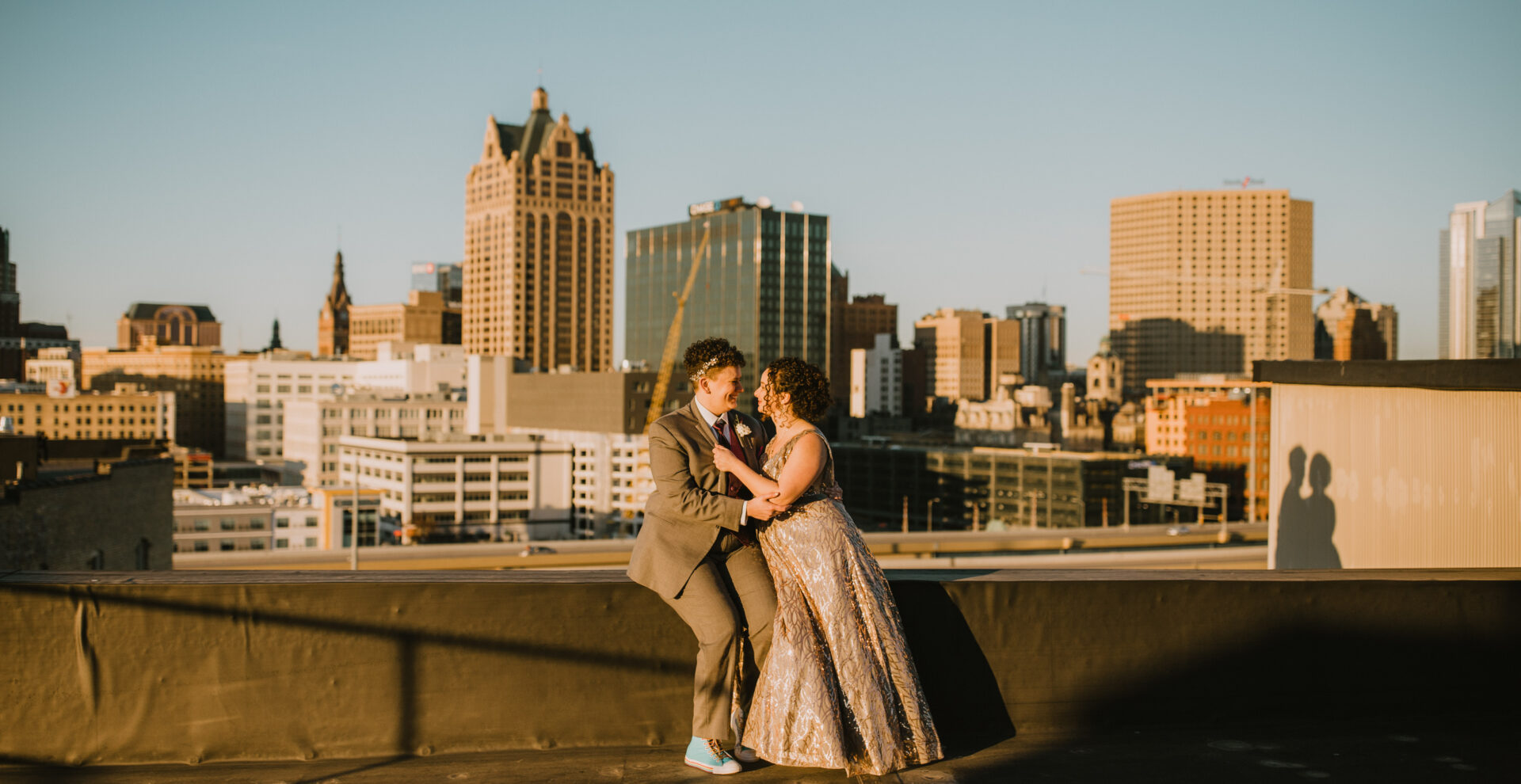 brides embracing while sitting on a rooftop at sunset with city view behind them