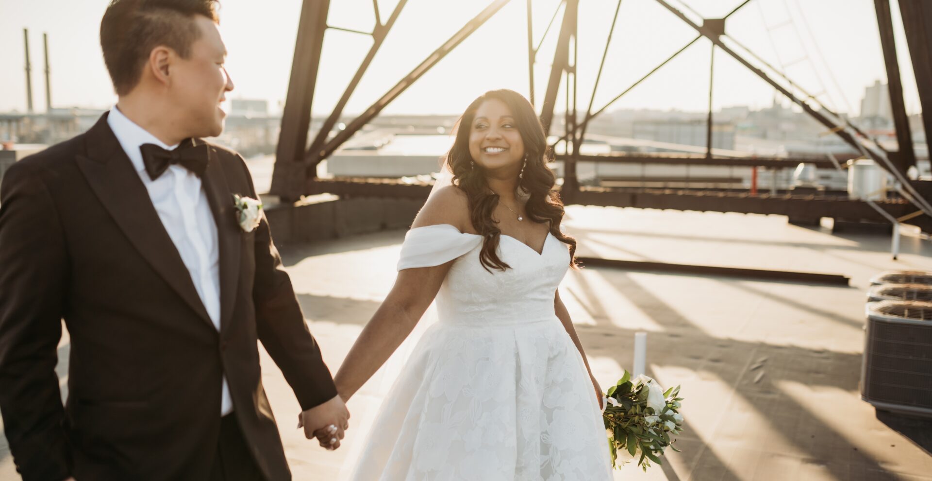 bride and groom holding hands and looking at each other while smiling on the rooftop at sunset