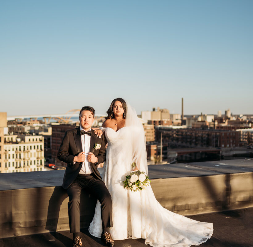 bride standing next to sitting groom on rooftop with a city view in the background