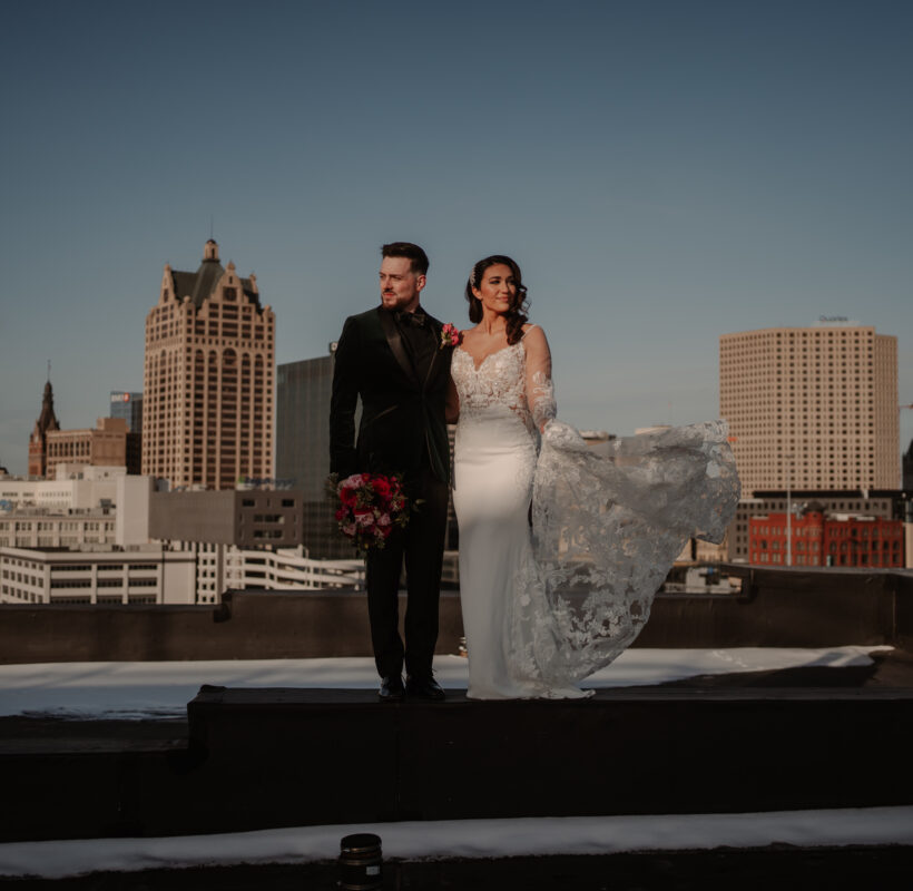 bride and groom standing on top of metal beam on rooftop at sunset with a vity view behind them